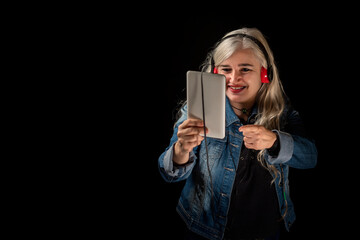 Happy gray haired woman in blue denim jacket making video conference with tablet and red headphones, pointing finger at electronic device
