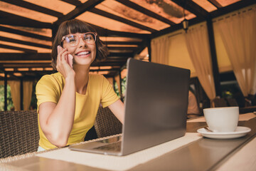 Photo of shiny positive lady dressed yellow t-shirt glasses sitting cafeteria drinking coffee talking modern gadget outdoors urban park