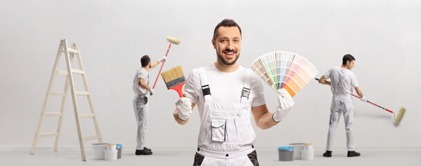 House painter holding a color swatch palette and a brush and other painters painting a wall - obrazy, fototapety, plakaty