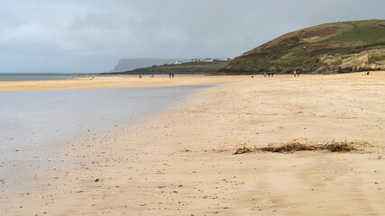 People and dogs strolling on a sandy bay in North Cornwall on a breezy November day lit by a shaft of sunlight. Trebetherick Point in the middle distance.