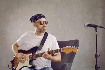 Obraz premium Cool man rocker in glasses play guitar record song on microphone in studio. Focused male musician or singer with musical instrument compose create music. Hobby, entertainment industry concept.