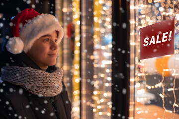 Boy in Santas hat looking and dreaming in illuminated shop window. Christmas holidays sales concept