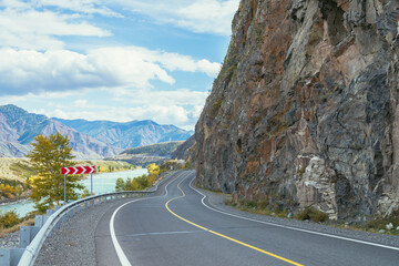Vivid autumn landscape with mountain highway along big mountain river in sunshine. Bright alpine scenery with wide turquoise river and mountain road in autumn colors. Highway in mountains in fall time