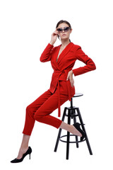 High fashion photo of a beautiful elegant young woman in a pretty red suit, jacket, pants, trousers and sunglasses on white background. Slim figure. Studio Shot. Businesswoman sitting on a black chair