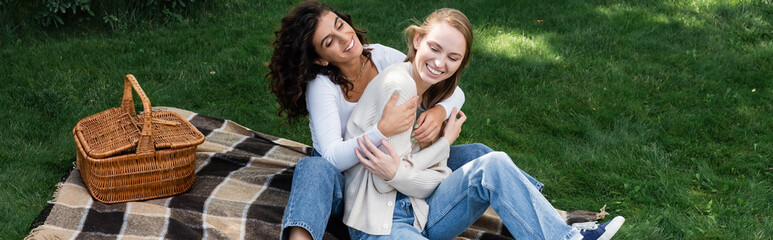 joyful lesbian women hugging on checkered blanket near basket, banner