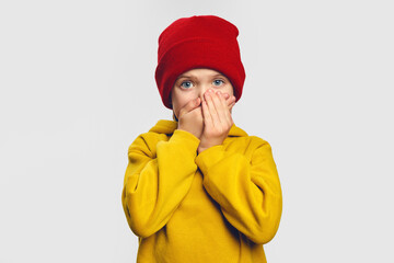 Terrified cute little girl covers mouth with great fear, tries not to cry, has scared shocked expression, wears yellow hoodie and red hat 
