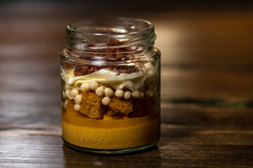 Dessert in a glass tub filled with cream cheese and caramel with white chocolate balls covered with dark chocolate and a crunchy biscuit base