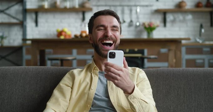 Relaxed young man sitting on sofa in living room, surfing internet on smartphone, getting online lottery win notification, celebrating success indoors, good luck achievement concept.