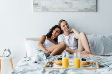 Obraz na płótnie Canvas happy young woman leaning on girlfriend near tray with breakfast on bed