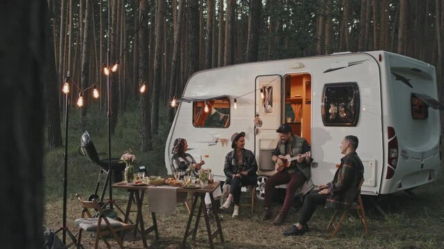 Slowmo shot of man sitting before cozy white campervan parked in forest on summer evening and playing song on ukulele for his happy friends