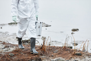 Low section shot of unrecognizable person wearing hazmat suit walking by water at ecological...