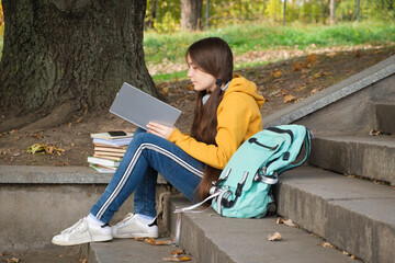 A girl of 13 years old sits on the stairs and reads a book in the park, goes to school exams.