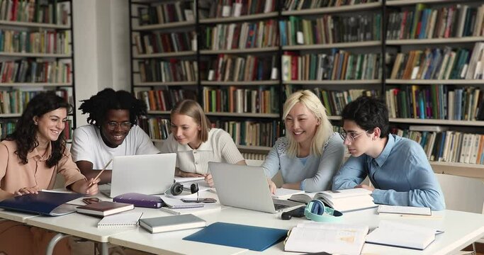 Group of multi ethnic positive students using laptop preparing for high school exams sit at table in library. Generation Z and modern tech usage for gaining new knowledge, e-learning or fun concept