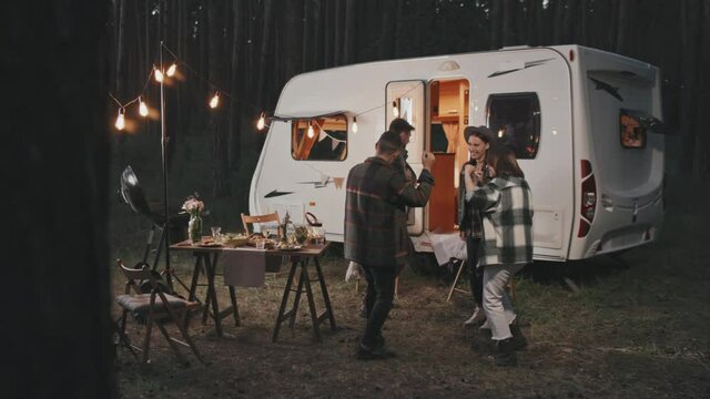 Slowmo shot of happy young people dancing together at cozy campground on summer evening. White campervan parked in background