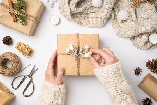 First person top view photo of female hands in sweater decorating craft paper giftbox with twine and snow branch scarf christmas tree balls and handicraft tools on isolated white background