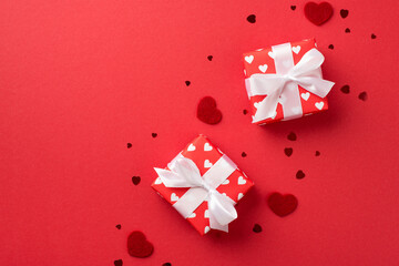 Top view photo of valentine's day decorations gift boxes in red wrapping paper with pattern of hearts and white ribbon bows decorative hearts and confetti on isolated red background with copyspace