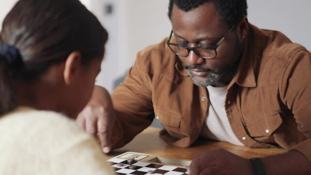 Cheerful African father and daughter playing checkers at home