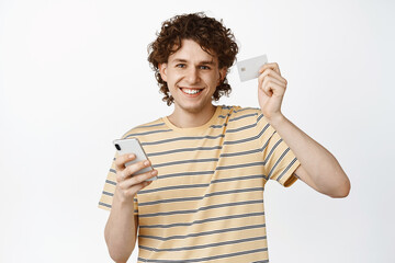 Handsome smiling curly guy showing credit card and smartphone, concept of online shopping, white background
