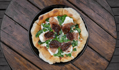 Top view of an arugula pizza with burrata cheese, sun dried tomatoes and cured raw ham, served in the pizza pan on the wooden table.