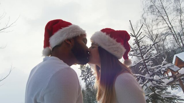 Christmas spirit. Young couple with santa hats kissing while holding heart shape framing the sun on the snowy mountain. High quality 4k footage