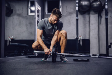 Naklejka na ściany i meble Working out in the gym with no other people, individual training. A shot of a handsome man in a grey shirt and shorts setting up equipment for training in a modern sports center. Fitness challenge