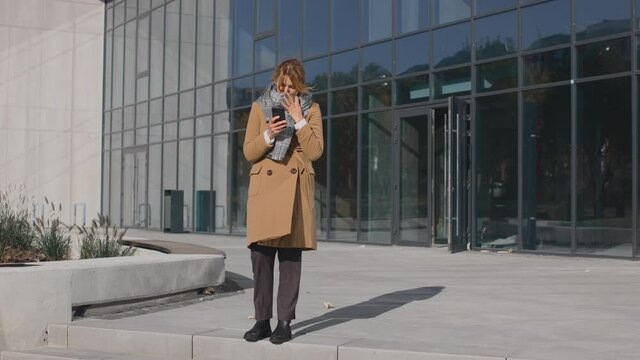 Full lngth of the caucasian woman coughing hard on the street in autumn while using her smartphone. Office building behind her