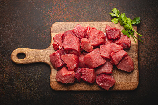 Raw beef meat chopped in cubes with bunch of fresh parsley, garlic, salt and pepper on wooden cutting board for cooking stew or meat dish on brown dark stone concrete background top view flat lay