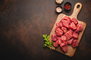 Raw beef meat chopped in cubes with bunch of fresh parsley on wooden cutting board for cooking stew or other meat dish on brown dark stone concrete background top view flat lay space for text - 472290141