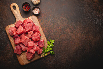 Raw beef meat chopped in cubes with bunch of fresh parsley on wooden cutting board for cooking stew or other meat dish on brown dark stone concrete background top view flat lay space for text