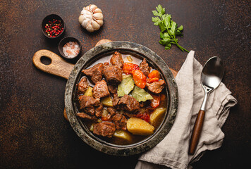 Delicious beef meat stew dish with potatoes, carrot and gravy in rustic metal bowl with spoon, bunch of fresh parsley, garlic cloves, spices on brown concrete background top view