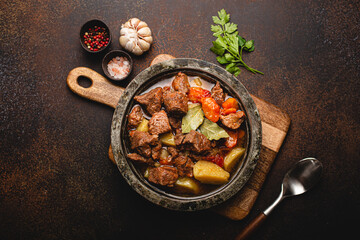 Delicious beef meat stew dish with potatoes, carrot and gravy in rustic metal bowl with spoon, bunch of fresh parsley, garlic cloves, spices on brown concrete background top view