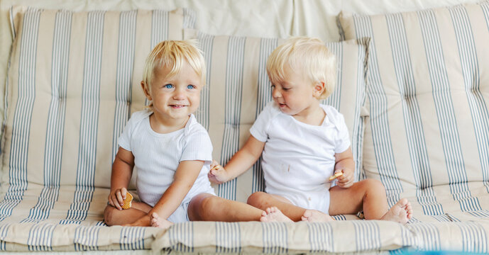 Babies eating cookies and playing around. 2 happy toddlers in white baby bodies holding a cake in their hand on a beige striped sofa One child smiles and looks away while the other looks at a relative