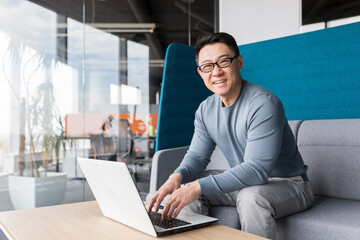 Asian businessman smiles and rejoices looking at camera, at work, working online with laptop, man in casual clothes, working at new startup in modern office