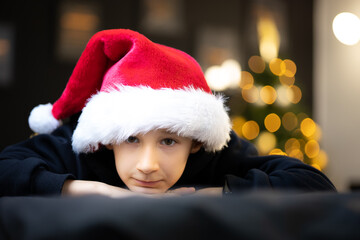 the boy in the Santa hat folded his chin on the sofa and looks at the camera against the background of Christmas tree toys, he is a little sad