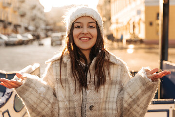 Cheerful fair-skinned young lady looks into camera smiling broadly on street of European city. Brown-eyed brunette spreads her arms to sides, wearing hat and coat. Rest time concept.