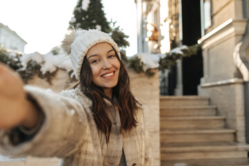 Cheerful caucasian young girl with toothy smile makes cute selfie against backdrop of European winter city. Brunette is in good mood, hat and jacket. Snow winter beauty concept.