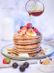 Selective focus of a healthy banana and oatmeal pancakes with fresh strawberries ,red grape, walnuts and grains with a pot of syrup pouring over 