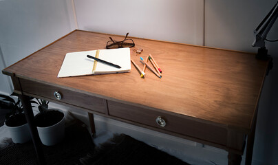 Close-up from a high angle of a white notepad next to a few wooden colorful pencils on a oak desk with a warm light over them.