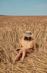 Young woman barefoot in linen clothes and a hat on a background of dry grass. Romantic girl in a hat sits on a meadow, relaxed alone in nature. View from above.