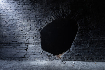 Dry dark brickwork tunnel of an old drainage collector of the 19th century with light coming from the street on the top left. Old brick wall with a hole in the center.