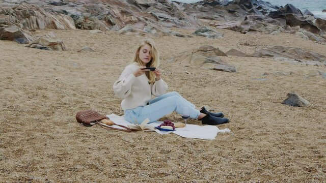 Young blonde woman, blogger, influencer, is sitting on blanket on beach, having small picnic and taking photos on her gadget, creating content to inspire subscribers, Slow motion.