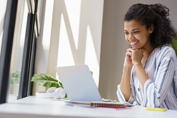 Beautiful smiling African American woman freelancer looking at digital screen working from home. Happy student using laptop computer, studying, learning something. Online education concept