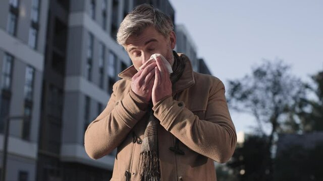 Sick Caucasian Senior Man Sneezing While Standing Outside Office Building. Portrait Of Ill Male Feeling Unwell And Coughing Outdoors While Using Napkin