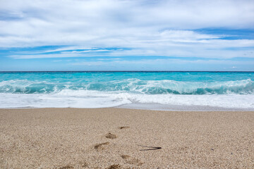 Fototapeta na wymiar Azure vibrant waves with blue sky on sunny coast of Greek island. Sandy beach with footprints in Greece. Summer nature vacation travel to Ionian Sea