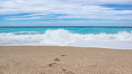 Fototapeta na wymiar Azure vibrant waves hitting shore with blue sky on sunny coast of Greek island. Sandy beach with footprints in Greece. Summer travel to Ionian Sea