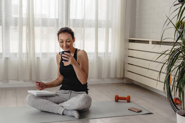 Young woman writing in her notebook, drinking coffee. Sitting at home by the window on a yoga mat after exercise. Personal growth resolutions
