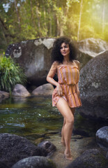 A young athletic sexual woman with black curly hairs  dressed in an orange short dress is posing in the tropical creek. Portrait Photography. -Image.