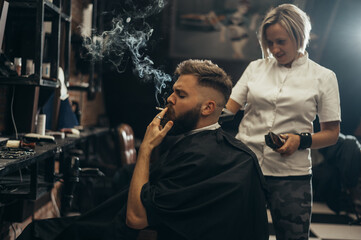 Young bearded man getting haircut by hairdresser and smoking a cigarette