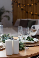 Round table decorated for Christmas and New Year. Bed in the background. Christmas tree twigs on the table. Plates and wine glasses on the table. Kitchen set and flowers on the background.