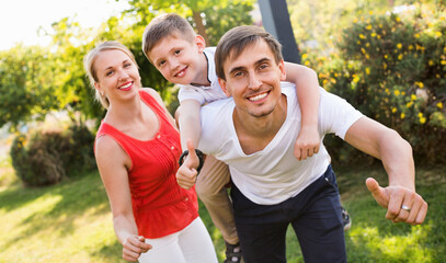 Portrait of positive smiling family with boy sitting on father's back hugging each other in park